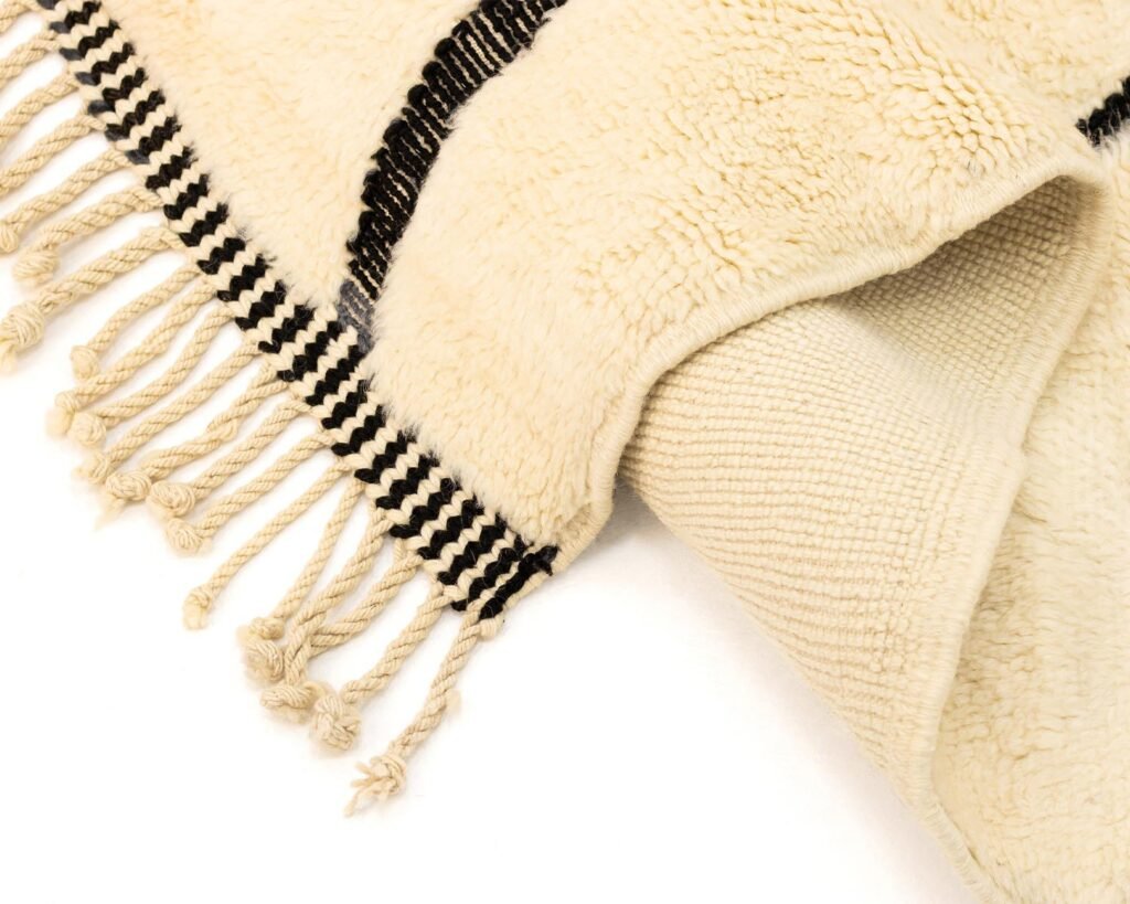Close-up of a beige woven luxury rug with black stripes and fringed edges on a white background.