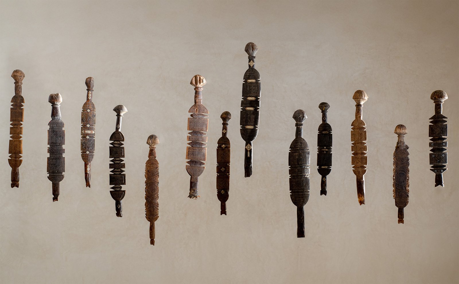 Eleven intricately carved wooden Berber Tent Pegs, sculptures of varying designs are mounted vertically on a plain wall.