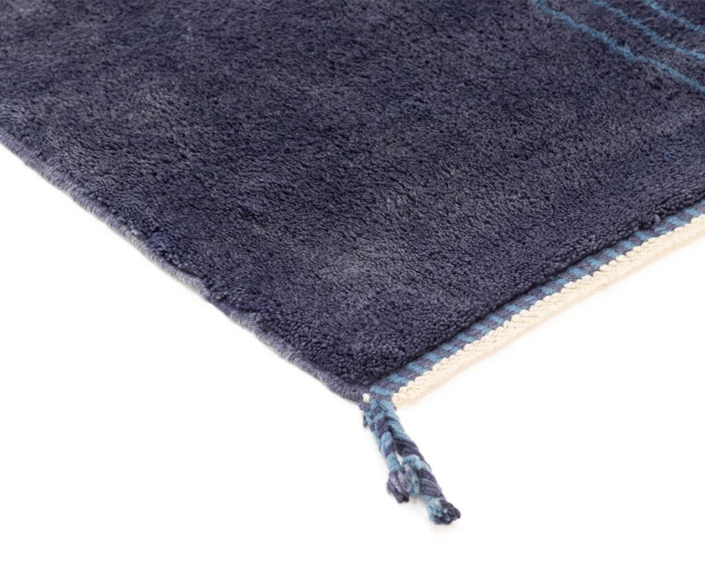Close-up of a dark blue textured rug with fringe tassels on the corner, placed on a white background.