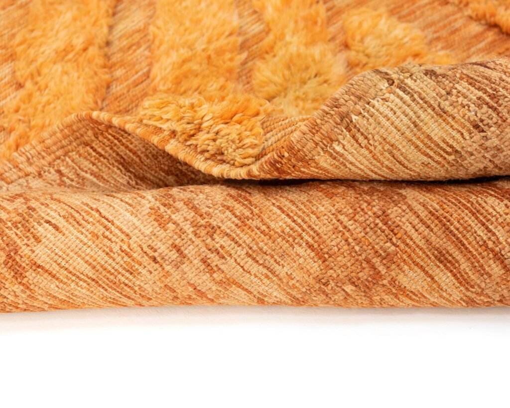 Close-up of a folded rug with a textured orange surface and a visible fluffy underside, displayed on a white background.