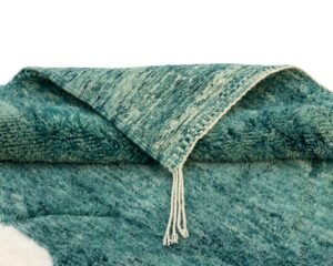 A folded teal and white textured rug with fringed edges against a white background evokes the intricate craftsmanship of artisanal rugs, offering a touch of elegance reminiscent of authentic Moroccan designs.