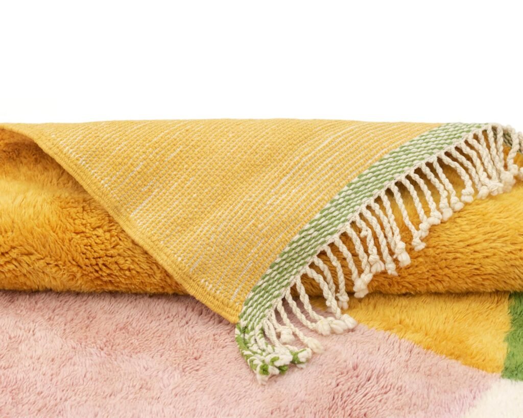 A close-up of a folded, multicolored fuzzy Rug with a yellow woven section featuring green and white fringe.