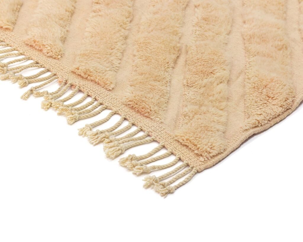 Close-up of a beige textured rug corner with fringe tassels, showcasing a soft, plush material and diagonal striped pattern.