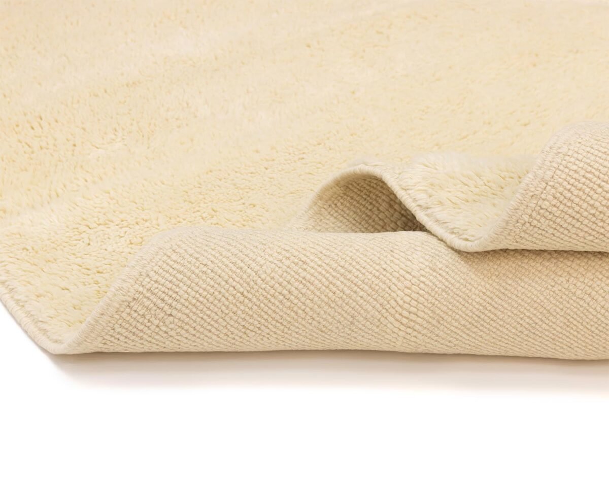 Close-up of a luxurious, beige shaggy rug with a folded corner revealing its textured underside. A hint of sophistication is infused in every strand, offering both comfort and elegance to any space.