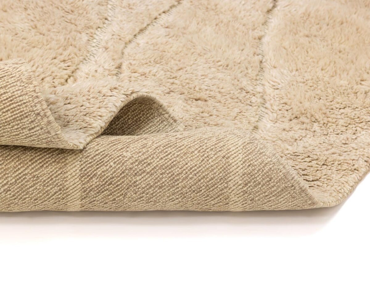 Close-up of a beige, textured luxury rug with one corner folded over, revealing its woven underside.