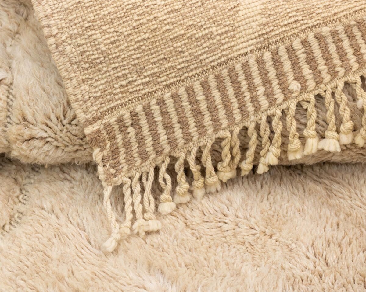 Close-up of a beige hand-knotted rug with fringed edges lying on a fluffy, light brown carpet.