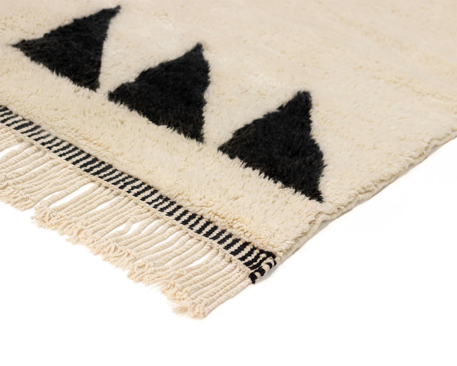 Corner of a hand-knotted, cream-colored rug with black triangle patterns and fringe edges, showcasing exquisite handmade home decor.