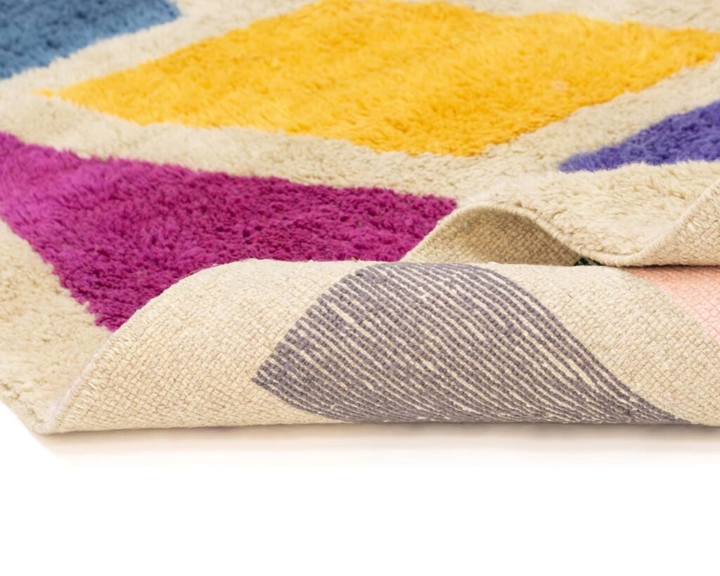 A colorful hand-knotted rug with purple, yellow, and blue diamond patterns is partially folded at the corner, revealing a textured underside. This piece of handmade home decor adds a touch of elegance to any room.