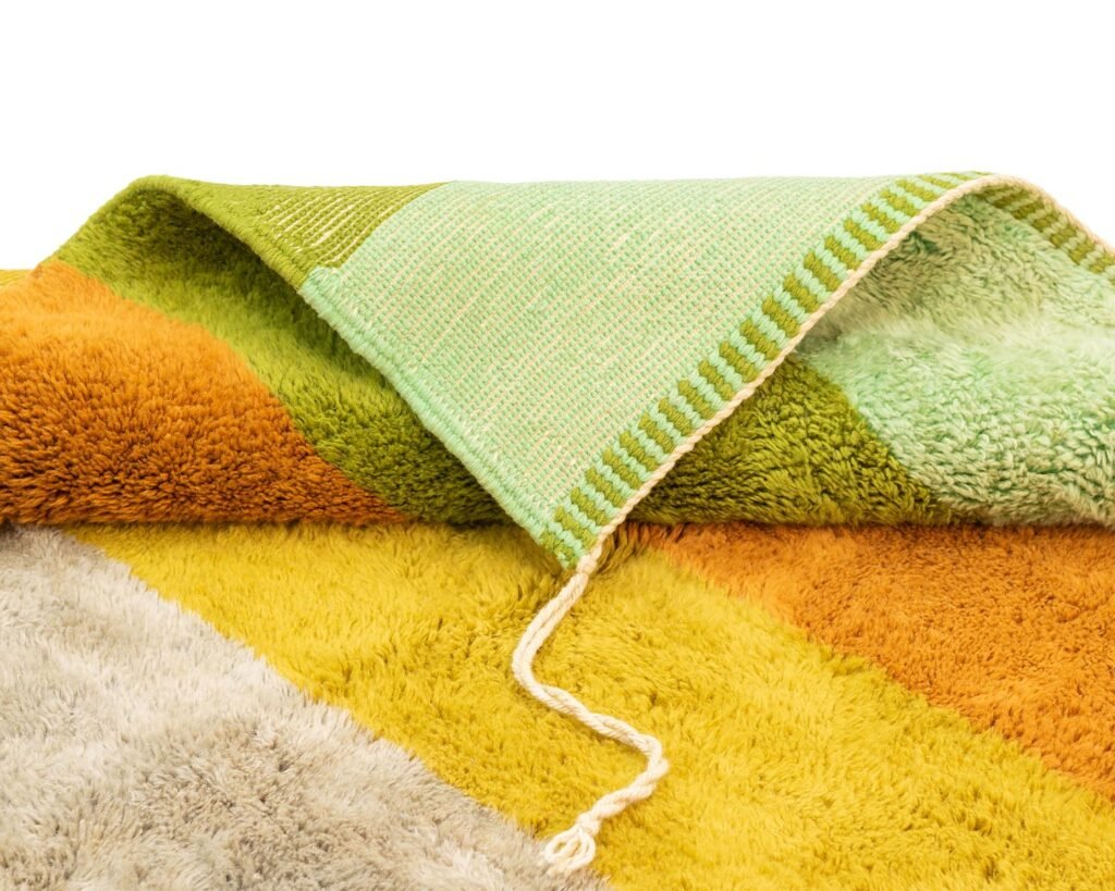 A close-up of a colorful, folded-up rug showing a mix of textured and flat patterns in shades of green, yellow, orange, and beige with a white fringe and a loose thread.