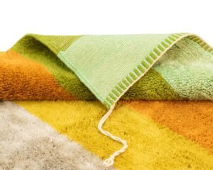 A close-up of a colorful, folded-up rug showing a mix of textured and flat patterns in shades of green, yellow, orange, and beige with a white fringe and a loose thread.