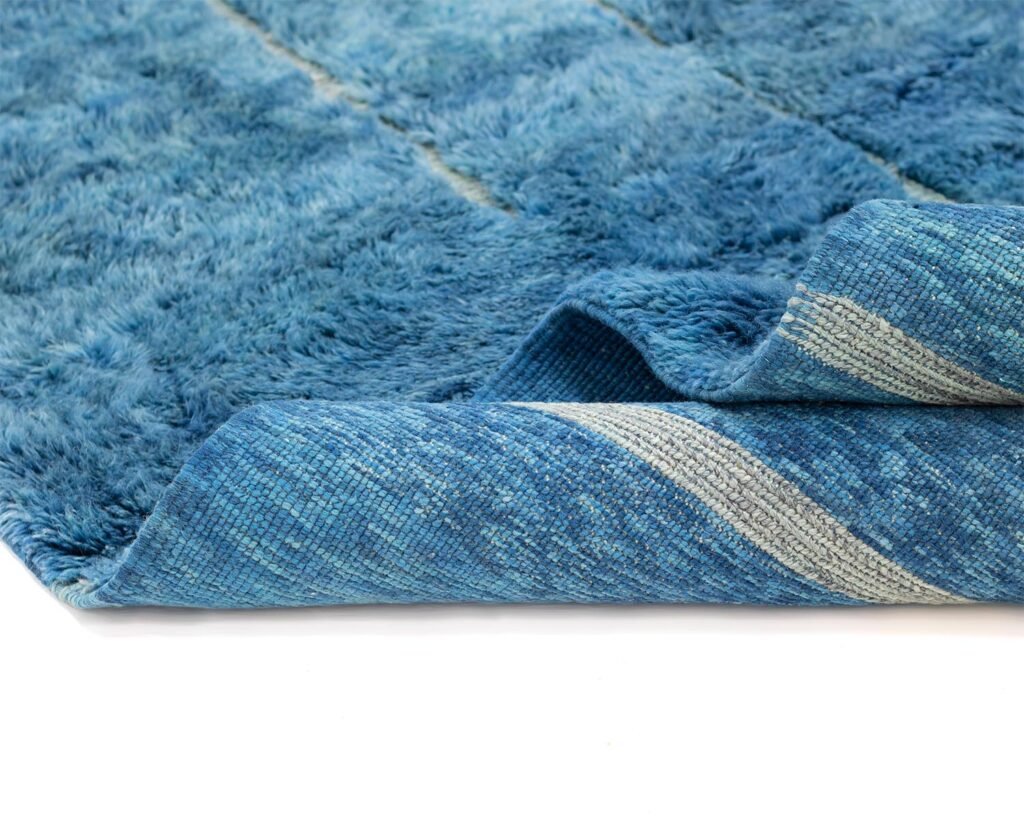 Hand knotted blue textured rug with a folded corner revealing a striped underside, set against a white background.