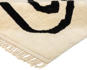 Close-up of a beige Moroccan rug corner with black abstract patterns and fringed edges, showcasing exquisite craftsmanship.