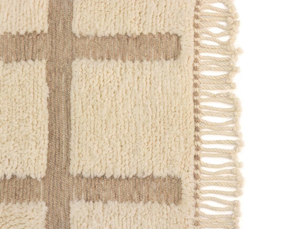 Close-up of a beige woven rug featuring rectangular patterns and fringed edges on the right side, adding a cozy touch to your room.