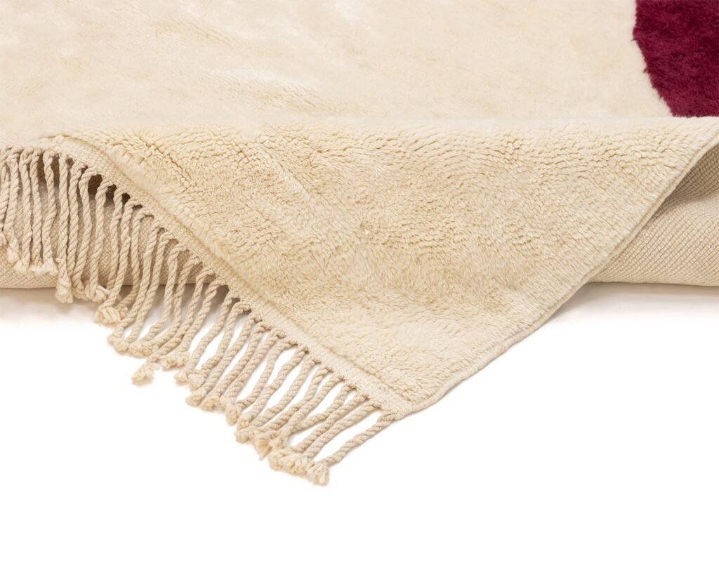 A folded cream-colored luxury rug reveals its tassel edge and intricate texture on a white background, showcasing the elegance of handmade home decor.