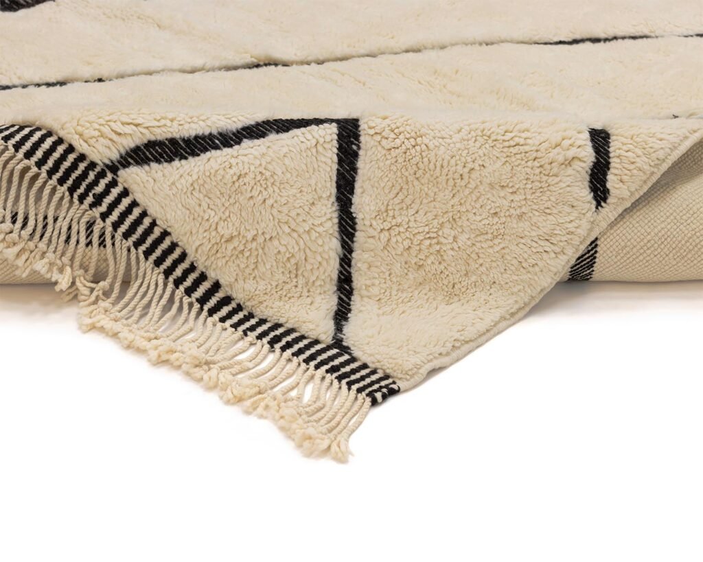 Close-up of a rug with black geometric lines and fringe on the edge, partially folded over at the corner. The intricate design adds a touch of elegance to any room.