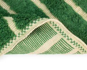 Close-up of a green and cream striped textured fabric with fringed edges and visible stitching detail, reminiscent of hand-knotted Moroccan rugs.