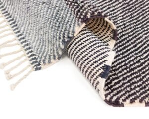 A black and white Moroccan rug with fringed edges is partially rolled over, unveiling a striped pattern on the underside.