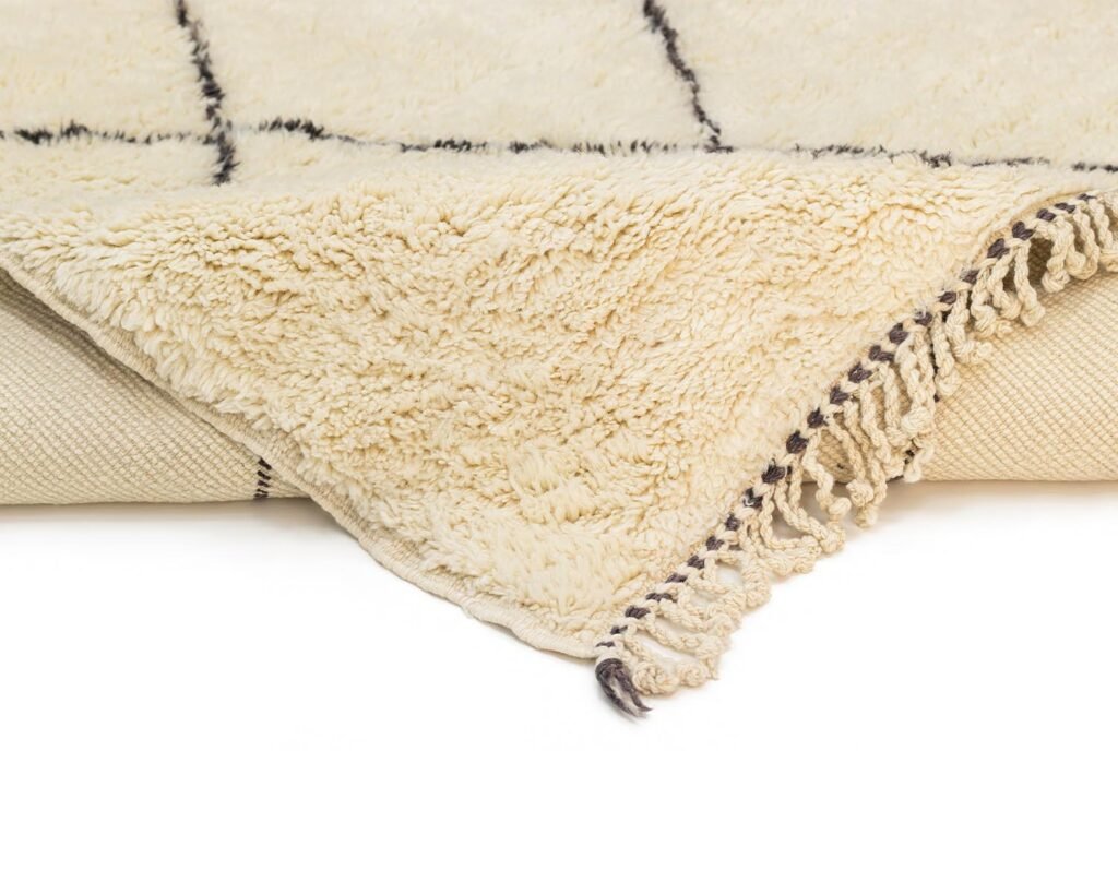 Close-up of a corner of a cream-colored, hand-knotted Moroccan rug with fringe edges, laid on a flat surface. Black lines form a simple grid pattern.
