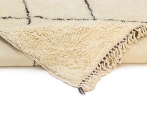 Close-up of a corner of a cream-colored, hand-knotted Moroccan rug with fringe edges, laid on a flat surface. Black lines form a simple grid pattern.