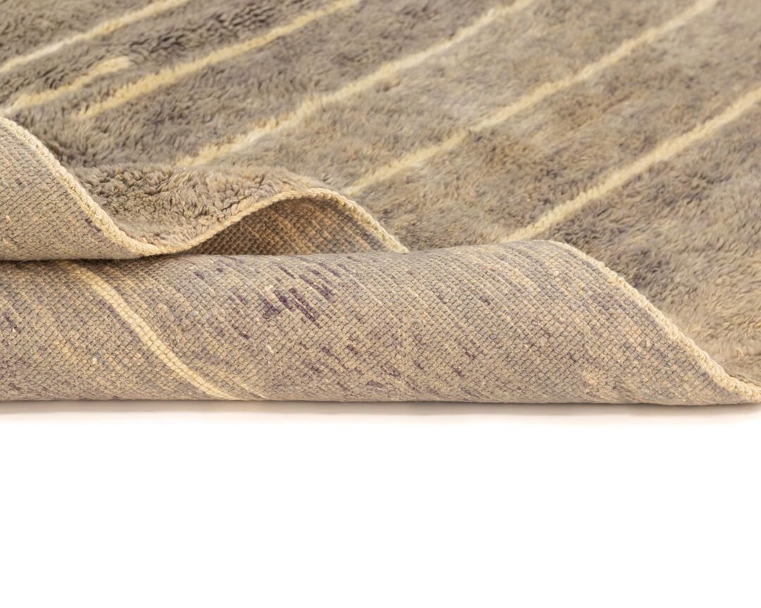 A close-up of a beige, textured rug with a striped pattern and intricate detail. One corner is folded over, revealing the woven underside for a contrasting view. The craftsmanship highlights its elegant design and quality materials.