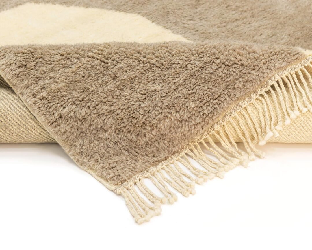 A beige and cream textured rug with fringe on one edge lies partially folded, revealing a lighter underside. This cozy addition enhances any rooms aesthetic with its warm tones and artisanal design.