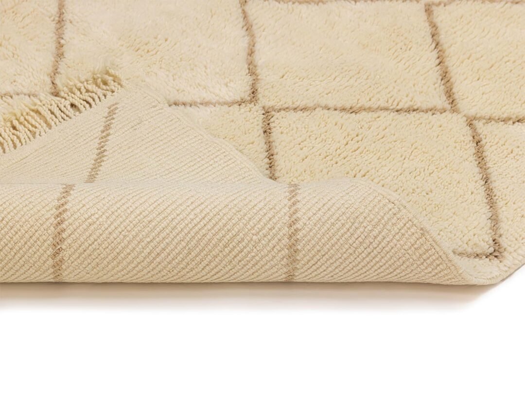 A beige luxury bath mat with a grid pattern is partially folded to reveal its fluffy top and textured underside, reminiscent of a Moroccan rug.