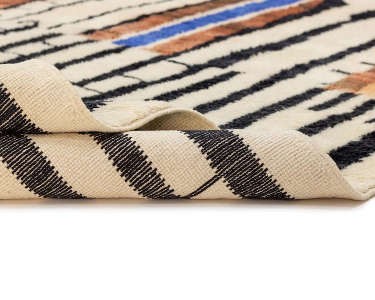 Close-up of a folded, patterned rug on a white surface. The rug boasts black stripes interwoven with earth-toned patterns and hints of blue on a cream background, evoking an artistic touch.