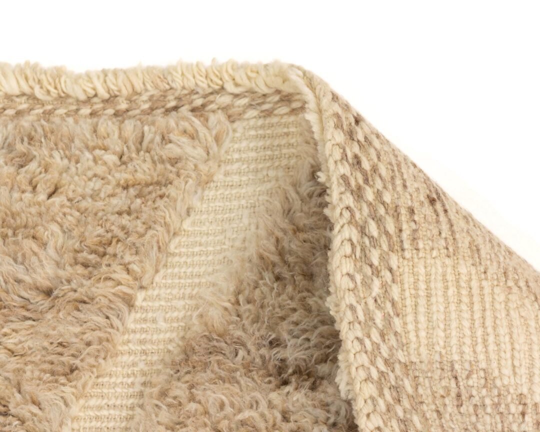 A close-up of a corner of a beige textured rug reveals its intricate woven pattern beneath, contrasted by the soft, fuzzy surface on one side. The folded section highlights the craftsmanship and adds depth to its elegant design.