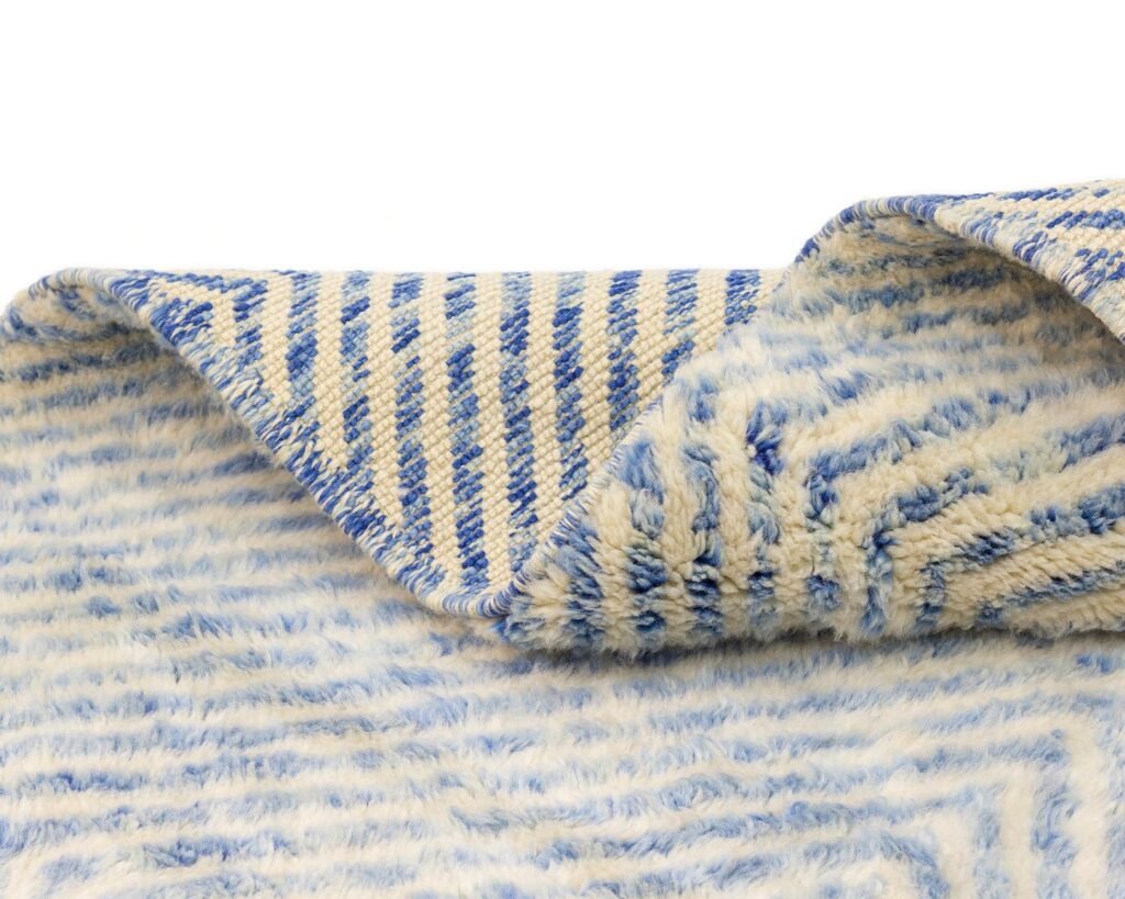 Close-up of a textured fabric with a geometric pattern in blue and white, reminiscent of luxury rugs. The fabric is slightly folded at the corner, adding a touch of elegance to this piece of handmade home decor.