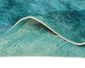 A close-up of a teal shaggy rug reveals its intricate design, with a corner flipped to uncover the detailed underside.