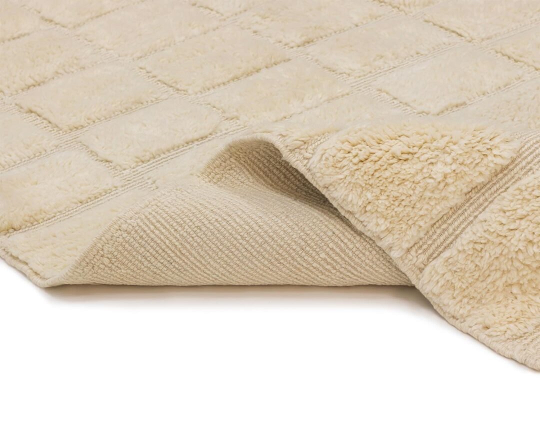 A luxurious beige checkered rug with a textured surface is partly folded to reveal its soft, fluffy underside, reminiscent of a hand-knotted Moroccan rug.