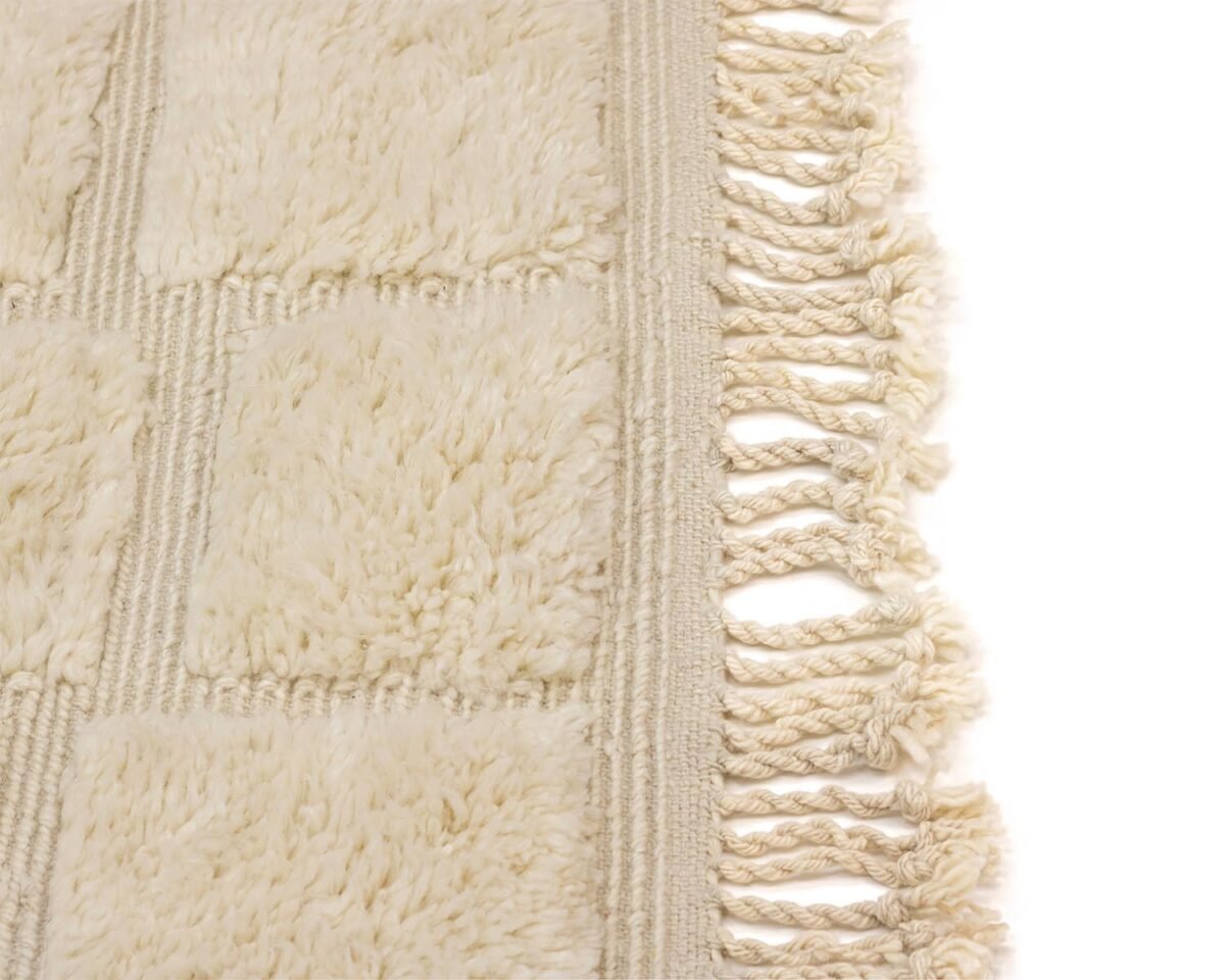 Close-up of a cream-colored, textured Moroccan rug with a checkered pattern and fringed edges on the right, exuding an air of luxury.