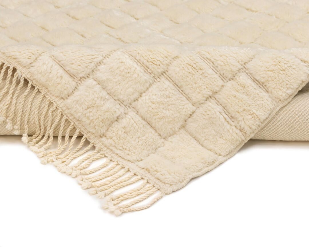 A beige, hand-knotted piece featuring a quilted pattern and tasseled edges is shown folded over, reminiscent of authentic Moroccan rugs.