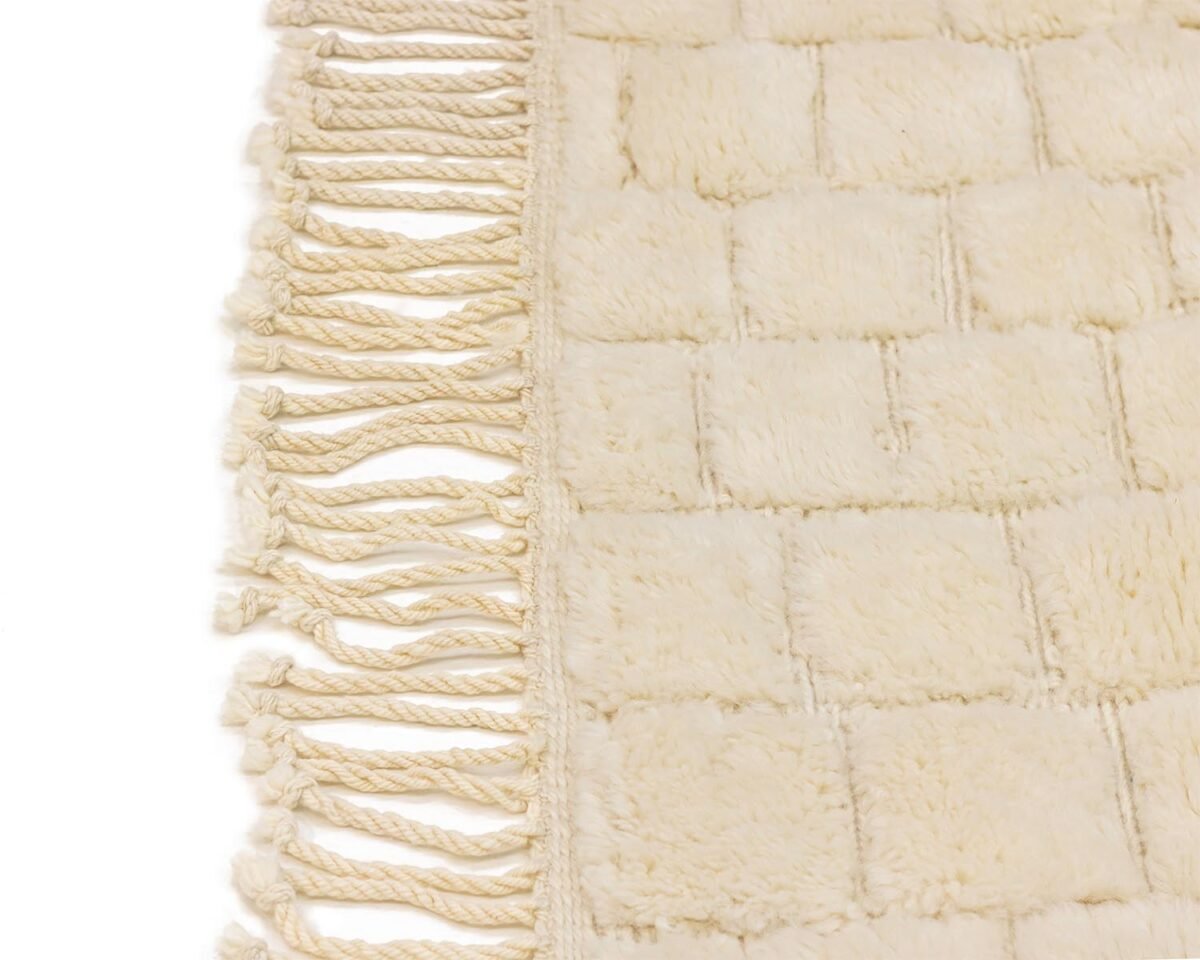 Close-up of an ivory shaggy rug with a braided fringe on the left, hand-knotted for added durability. The texture appears soft and plush with a grid pattern, highlighting the craftsmanship typical of Authentic Moroccan Rugs.