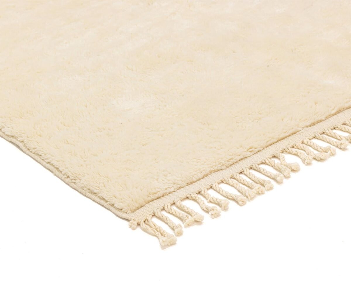 A corner of a beige, plush rug adorned with tassels rests elegantly on a white background, adding a touch of sophistication.