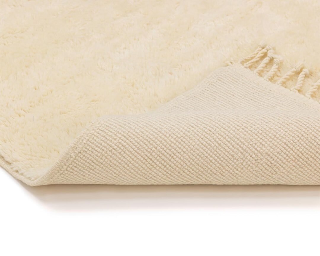 Close-up of a cream-colored rug folded at the corner to reveal its woven underside, with fringes elegantly draping along the edge.