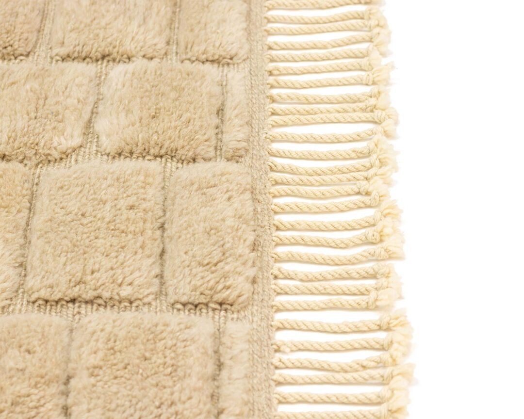 Close-up of a beige textured rug featuring a subtle square pattern and delicate fringe tassels along one edge, exuding warmth and style.