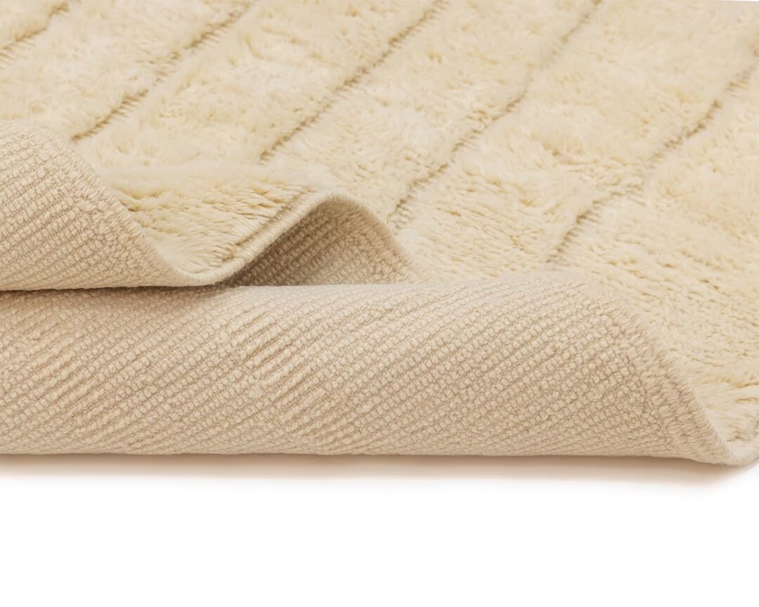 Close-up of a beige textured rug, partially folded to reveal a striped pattern on the underside and a plush surface on top. This beautiful piece adds warmth and style to any room, making it an ideal choice for cozy spaces.