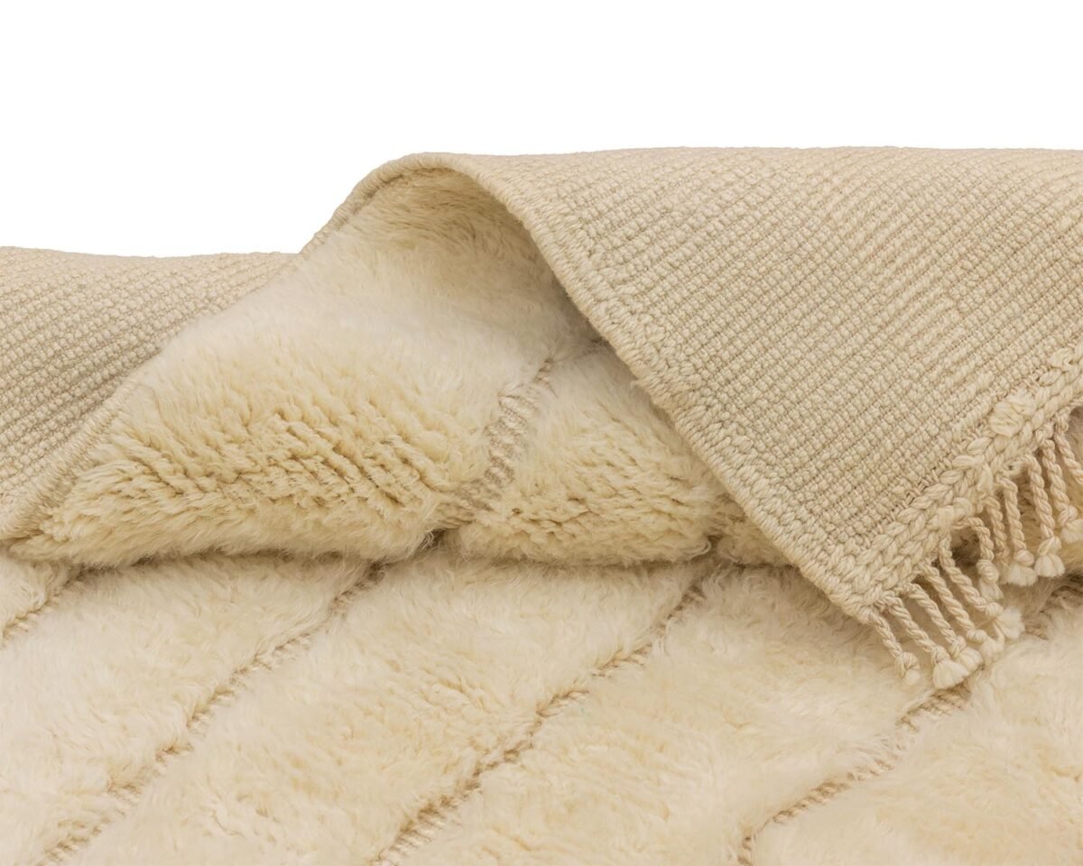 A beige, textured throw is folded to reveal its soft, fluffy underside and fringed edge, offering a warm and inviting touch.