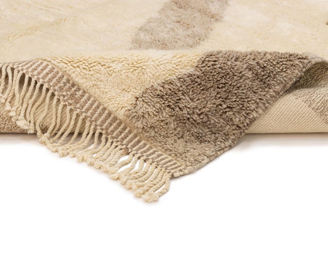 A beige and brown patterned rug with fringe on one corner is elegantly rolled to display both its intricate sides.