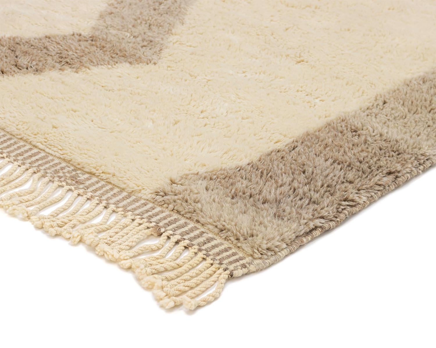 A close-up of a beige and tan shaggy rug corner with fringed tassels showcases its handmade charm. The texture appears soft and plush, perfect for adding a touch of cozy elegance to your home decor.