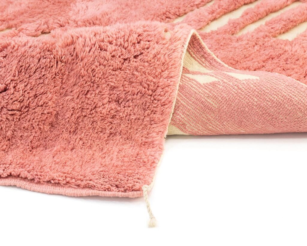 Close-up of a pink textured Moroccan rug with a corner slightly folded over, revealing the underside. A small string is visible near the edge, reflecting its exquisite hand-knotted craftsmanship.