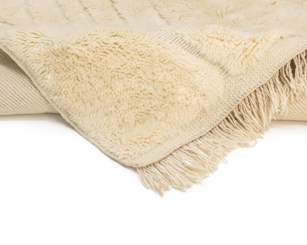 A detailed close-up showcases the cream-colored, fringed elegance of an authentic Moroccan rug, boasting a soft, textured surface. This hand-knotted piece reflects exquisite craftsmanship and artistry.