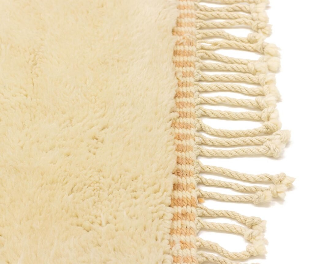 Close-up of a hand-knotted, beige fringed luxury rug partially captured in the frame, showcasing its fluffy texture and intricate fringe detail on one side.