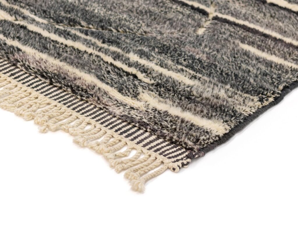 Close-up of a plush, gray and cream-colored rug with fringe on one edge. The rug showcases a striped pattern and soft surface.
