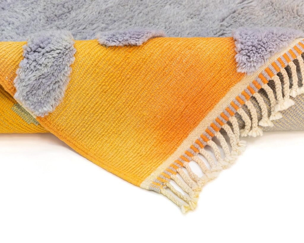 A yellow and orange, hand-knotted rug with fringe edges is partially flipped over a grey, fluffy textile surface.