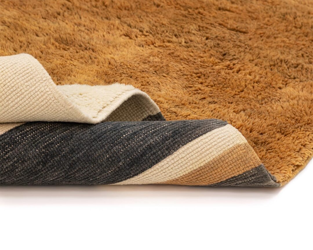 Close-up of a partially folded rug displaying its fluffy brown surface contrasted by a woven striped underside in black, beige, and gray.