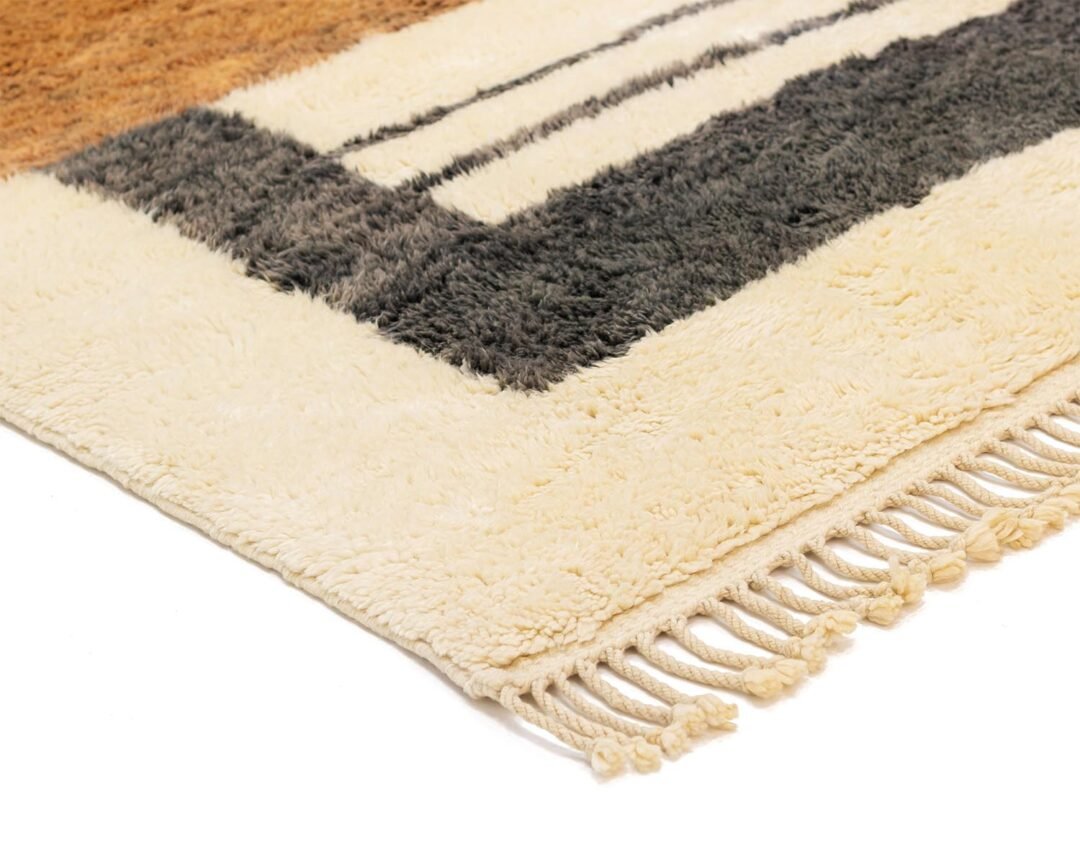 Close-up of a rectangular area rug featuring beige, black, and brown geometric patterns with fringed edges on one side, placed against a minimalist white background.