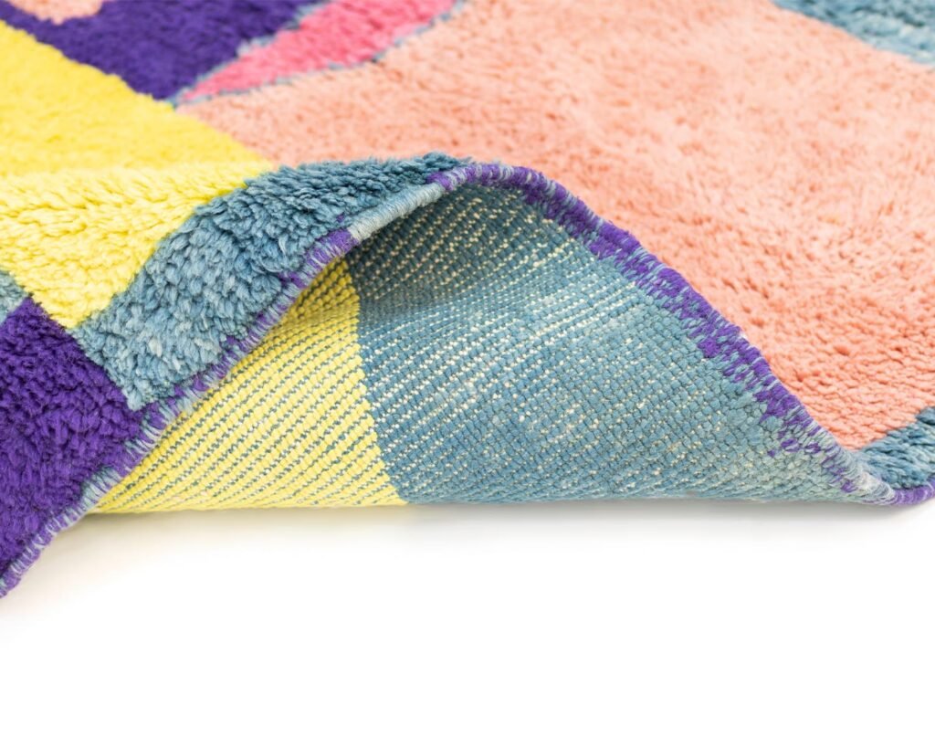 A vibrant, colorful, checkered rug showcases sections of pink, yellow, blue, and purple. The edge is partly folded to reveal its intricately woven underside.