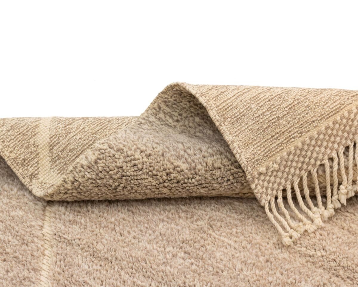 A luxurious beige woven rug with fringed tassels on one end, partially folded to showcase its texture and craftsmanship.